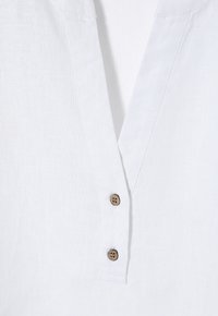 Hvid linned skjorte med v-hals design, prydet med to brune knapper ved halsudskæringen og en subtil stofstruktur.
