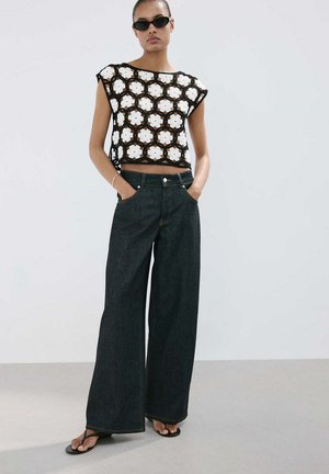 Mujer que lleva un top corto floral de ganchillo en blanco y negro, vaqueros anchos oscuros, gafas de sol negras y chanclas negras, de pie con las manos en los bolsillos.