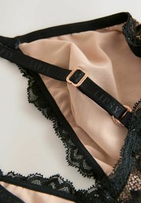 Spodné prádlo v telovom sieťovinovom materiáli s čiernymi čipkovými detailmi, s nastaviteľnými ramienkami a zlatou sponou označenou "LIPSY". Textúrovaná čipková aplikácia.