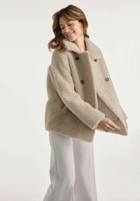 Kobieta uśmiechająca się, ubrana w beżowy, podwójnie zapinany futrzany płaszcz z dużym kołnierzem oraz białe spodnie z szerokimi nogawkami, na tle jasnego, jednolitego tła.