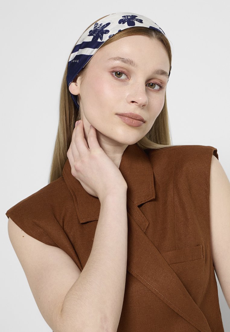 Giovane donna con lunghi capelli lisci che indossa un foulard floreale blu scuro e bianco e un blazer marrone senza maniche, mentre si tocca il collo.