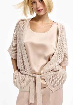 Blonde Frau, die ein hellrosa satinärmloses Oberteil und eine beige gestrickte offene Strickjacke mit einem Stoffgürtel trägt, der in der Taille gebunden ist.