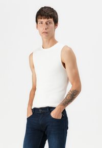 Fiatal férfi, rövid barna hajjal, fehér ujjatlan felsőt és sötétkék farmert viselve, a bal alkarján tetoválással, kezét a zsebében tartva.