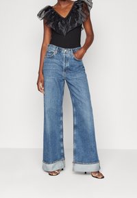 Kvinna i svart volanglös topp utan ärmar, vida blå jeans med uppvikta ben och svarta öppna klackskor, står med handen i fickan.