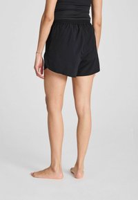 Zwarte sportshorts met een elastische tailleband, zijsplitten en een gladde textuur, gezien vanaf de achterkant met blote voeten zichtbaar.