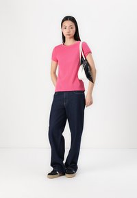 T-shirt rose à manches courtes, jeans larges bleu foncé, baskets noires avec semelles beiges, et un sac à main blanc avec une écharpe noire en accent.