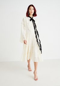 Rochie albă cu mânecă lungă, cu guler ruffled și fustă în straturi, împodobită cu puncte mici, accentuată cu un săculeț negru la gât.