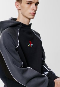 Ciemnoszara bluza z kapturem z wielokolorowym logo PlayStation na piersi, z kontrastującymi białymi szwami i ściągaczami.