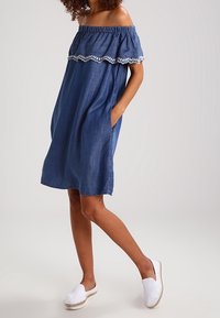 Mujer vestida con un vestido de mezclilla azul con hombros al descubierto y ribete bordado blanco, con las manos en los bolsillos y zapatos tipo espadrille blancos sin ataduras.
