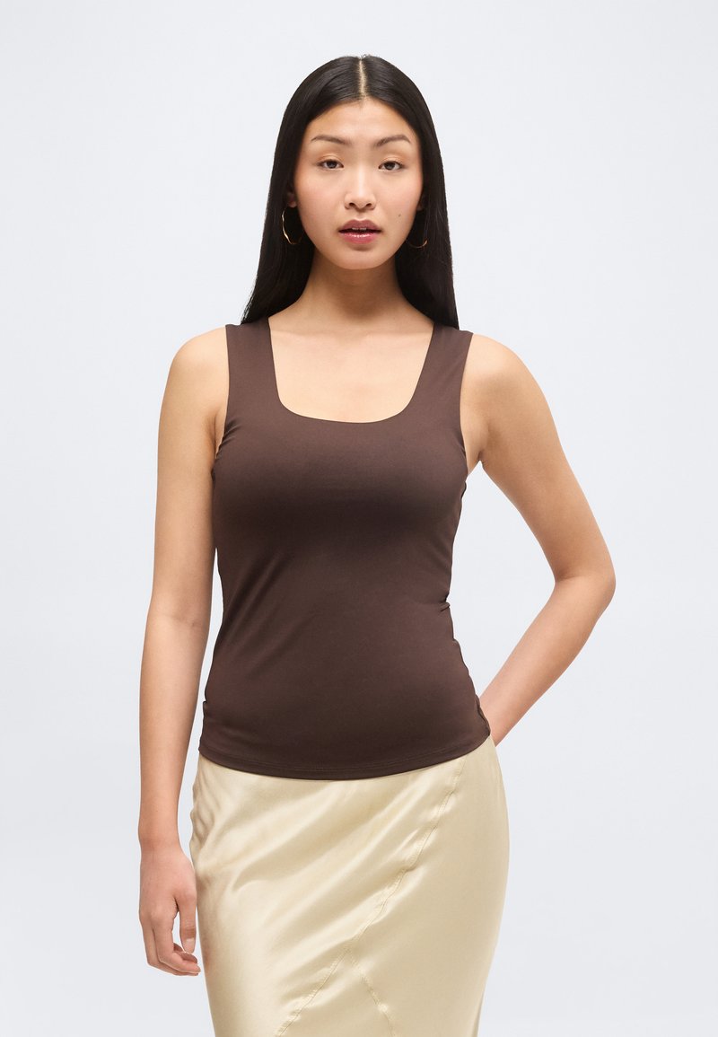 Femme aux longs cheveux noirs portant un haut marron sans manches et une jupe en satin crème, debout avec une main sur la hanche sur un fond uni.