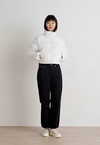 Vit puffjacka med hög krage, dragkedja fram och quiltad struktur, kombinerad med svarta vida jeans och vita sneakers.