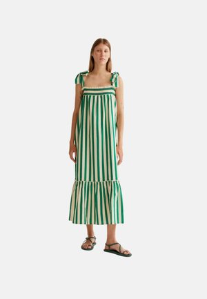 Mujer con un vestido midi sin mangas de rayas verticales verdes y blancas, con tirantes atados en los hombros y sandalias negras, de pie sobre un fondo blanco.