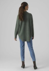 Oversized green sweater with a ribbed neckline, long sleeves, and a high-low hem, paired with light blue jeans and dark heeled boots.
