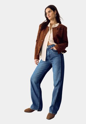 Femme portant une veste en daim marron, un chemisier crème, un jean à jambes larges bicolore et des chaussures en daim marron, posant avec la main dans la poche, regardant vers la gauche.