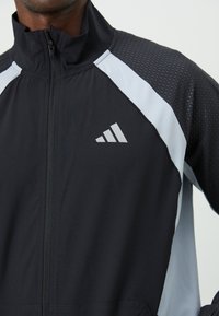Black zip-up jacket featuring light grey accents, textured shoulders, and a three-stripe logo on the left chest, made from lightweight fabric.