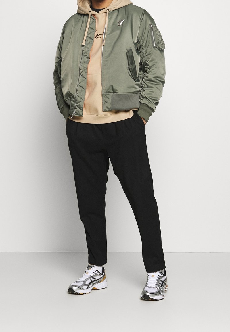Giacca bomber verde in satin con polsini a costine, felpa beige sotto, pantaloni neri affusolati e scarpe da corsa argento e oro.