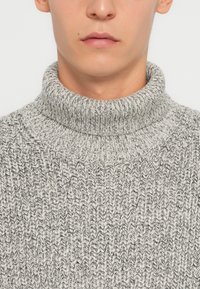 Maglione grigio a collo alto in maglia con un design testurizzato, caratterizzato da un collo alto e una vestibilità morbida. Il tessuto mostra fili chiari e scuri.