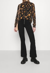 Blouse noire à motifs floraux orange, associée à un pantalon évasé noir à taille haute en côtes. Des bottes à plateforme noires complètent la tenue.