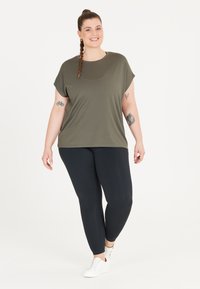 Olivgrön kortärmad topp i avslappnad passform, matchad med svarta leggings. Modellen bär vita sneakers och har synliga tatueringar.