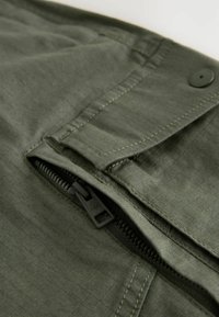 Gros plan sur un tissu vert avec une fermeture éclair noire partiellement ouverte et un rabat cousu maintenu par un bouton noir.