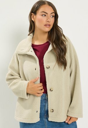 Kvinde med langt brunt hår iført en beige fleecejakke over en bourgognerød top og blå jeans, kigger til venstre.