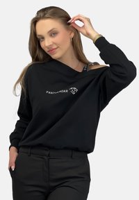 Czarna bluza z dekoltem opadającym na jedno ramię, z białym napisem "PANTHER ROAR" i prążkowanymi akcentami. W połączeniu z czarnymi spodniami.