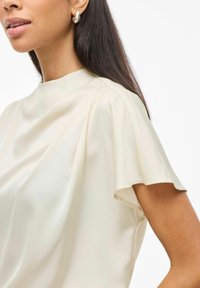 Satenasta ivory bluzica s visokim izrezom, kratkim rukavima in tekočim draperanjem; gladka tekstura, minimalističen dizajn in subtilen sijaj.