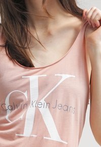 Ljusrosa ärmlös topp med bred halsringning och vitt "CK"-logotyp, med "Calvin Klein Jeans" i mindre text nedanför. Mjuk tygkänsla.