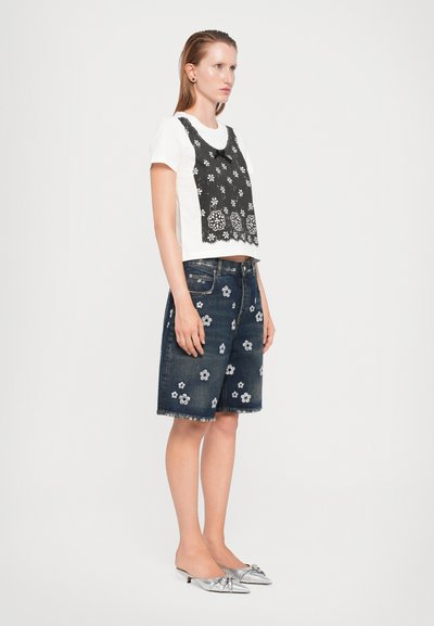 Witte t-shirt met een zwarte bloemenkant overlay, gecombineerd met donkere denim shorts met witte bloemenborduursels. Zilveren schoenen met een lage hak.