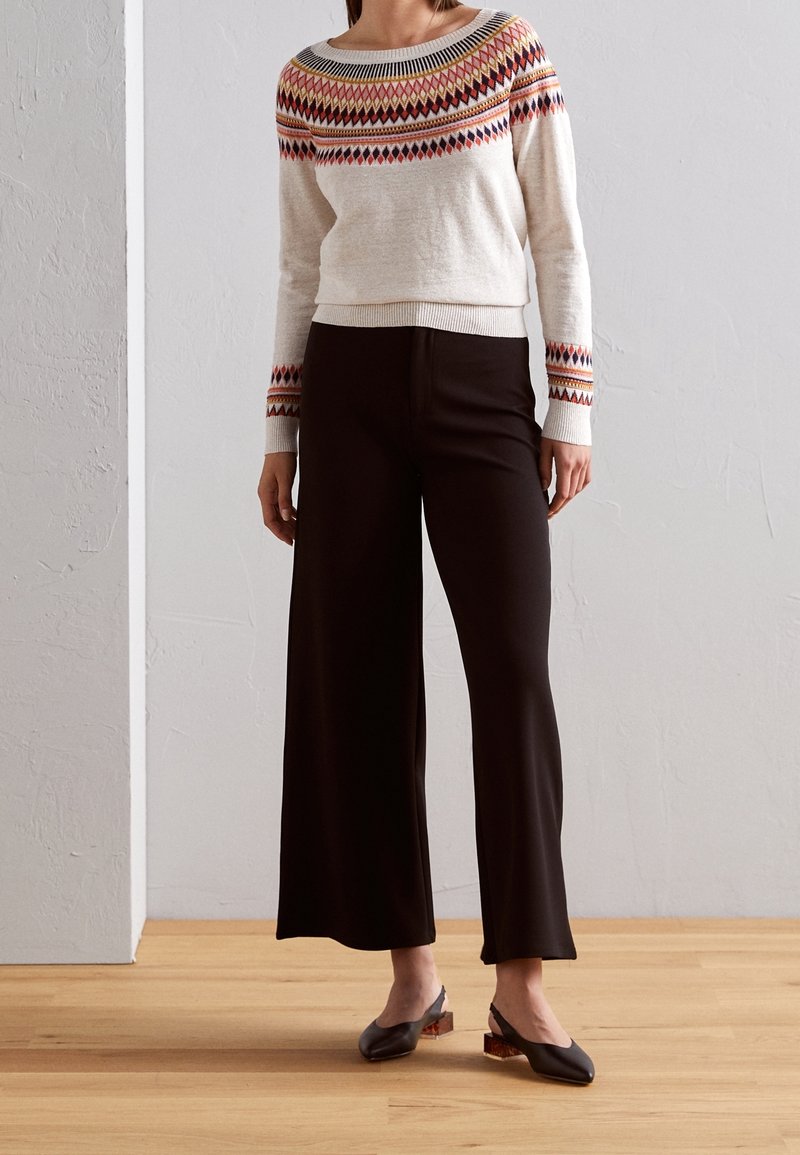 Femme portant un pull crème avec un motif losange multicolore et un pantalon large noir, debout sur un sol en bois devant un mur blanc.