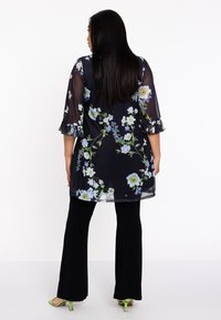 Doorschijnende zwarte tuniek met bloemenprint in blauw en groen, met ruches aan de mouwen en gecombineerd met zwarte flared broeken en groene hakken.