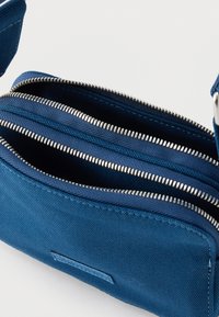 Μπλε υφασμάτινη τσάντα χιαστί με δύο ανοιχτά ασημένια φερμουάρ και ρυθμιζόμενο λουρί σε λευκό φόντο.