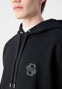Juodas džemperis su styginiu gaubtu, turintis tekstūruotą, siuvinėtą logotipą krūtinėje. Audinys turi lygią, patvarią išvaizdą.