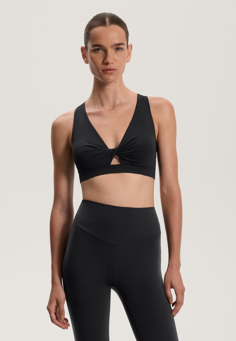 Sutien sport negru cu decolteu în V și detalii răsucite în față. Fabricat dintr-un material neted și elastic. Se asortează cu colanți negri cu talie înaltă.