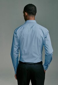 Hellblaues Langarmhemd mit Button-Down-Kragen, kombiniert mit schwarzen Anzughosen. Glattes Material, figurnahe Form und Knopfdetails an den Manschetten.