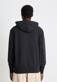 Person klädd i en svart huvtröja och ljusa beigefärgade byxor, stående vänd mot en enfärgad vit bakgrund med avslappnade händer vid sidorna.