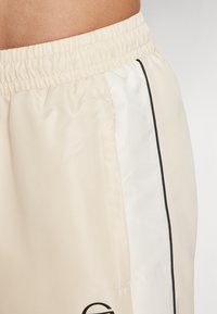 Krēma krāsas šorti ar elastīgu jostasvietu, kuriem ir balts sānu panelis un melna apdare. Gluda, viegla auduma struktūra ar maigu tekstūru.