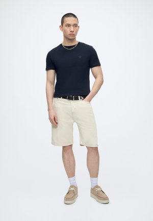 Tânăr purtând un tricou negru, pantaloni scurți bej, pantofi bej cu șosete albe, stând cu o mână în buzunar pe un fundal simplu.