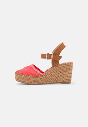 Sandał na koturnie z koralowym syntetycznym wierzchem, brązowym paskiem przy kostce i plecionym obcasem z juty. Posiada obszycie i detal z logo na wierzchu.