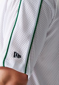 Camisa de malla blanca de manga corta con ribetes verdes a lo largo de la manga. Presenta un logotipo negro en la manga cerca del dobladillo.