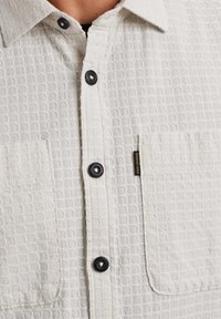 Camisa blanca texturizada con un patrón de panal, con botones negros y dos bolsillos en el pecho. Incluye una etiqueta de la marca en el lateral.