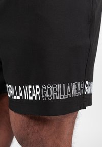 Crne kratke hlače od glatkog materijala s bijelim natpisom "GORILLA WEAR" duž rubova. Dizajn uključuje uski kroj i minimalne šavove.