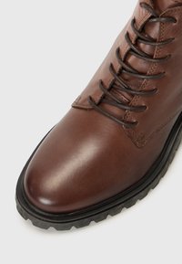 Botte en cuir marron avec un bout arrondi et des lacets. La semelle est robuste et présente des motifs de rainures distinctifs, le tout sur un fond blanc.
