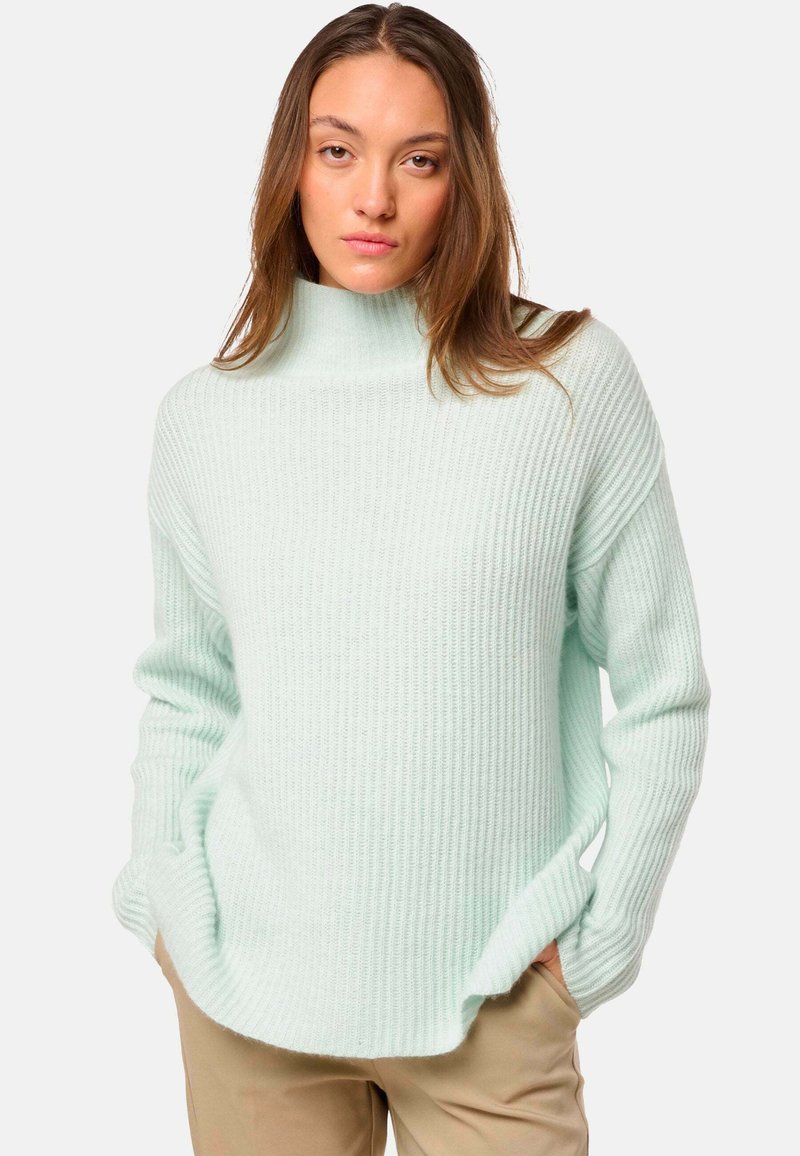 Jeune femme avec de longs cheveux bruns portant un pull col roulé à côtes bleu clair et un pantalon beige, les mains dans les poches.