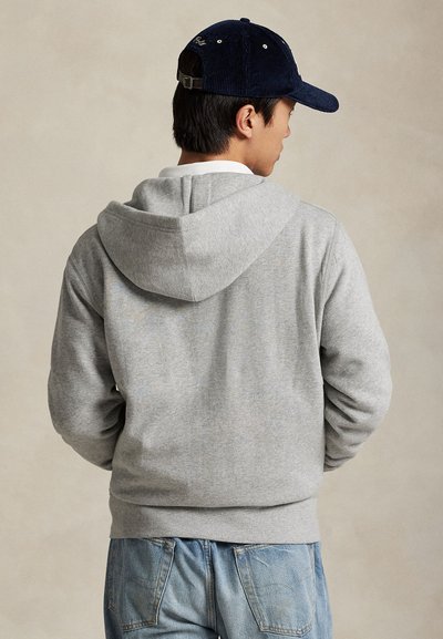 Mann trägt einen grauen Kapuzenpullover, eine blaue Kord-Baseballkappe und blaue Jeans, steht mit dem Rücken vor einem einfarbigen beigen Hintergrund.