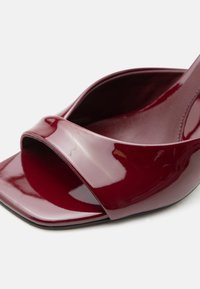 Γυαλιστερό μπουρντό ανοιχτό παπούτσι mule με φαρδιά λουρίδα και τετράγωνη μύτη σε απλό λευκό φόντο.