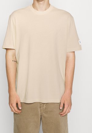 Mann trägt ein schlichtes, beiges Oversized-T-Shirt und hellbraune Hosen, mit einem kleinen Tattoo an der inneren linken Unterarmseite und einem Logo-Patch auf dem Ärmel.