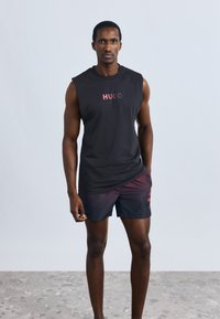 Črna majica brez rokavov z napisom "HUGO" v rdečih črkah. Kombinirana s črnimi in bordo kratkimi hlačami z neznatnim diagonalnim vzorcem.