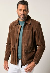 Hombre con barba que lleva una chaqueta de ante marrón sobre un suéter de color teal, de pie contra un fondo neutro, con las manos reposando en las caderas.