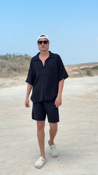 Ung man i svart skjorta och shorts, vit keps och skor, som går på en sandig väg med torra buskar och klar himmel i bakgrunden.